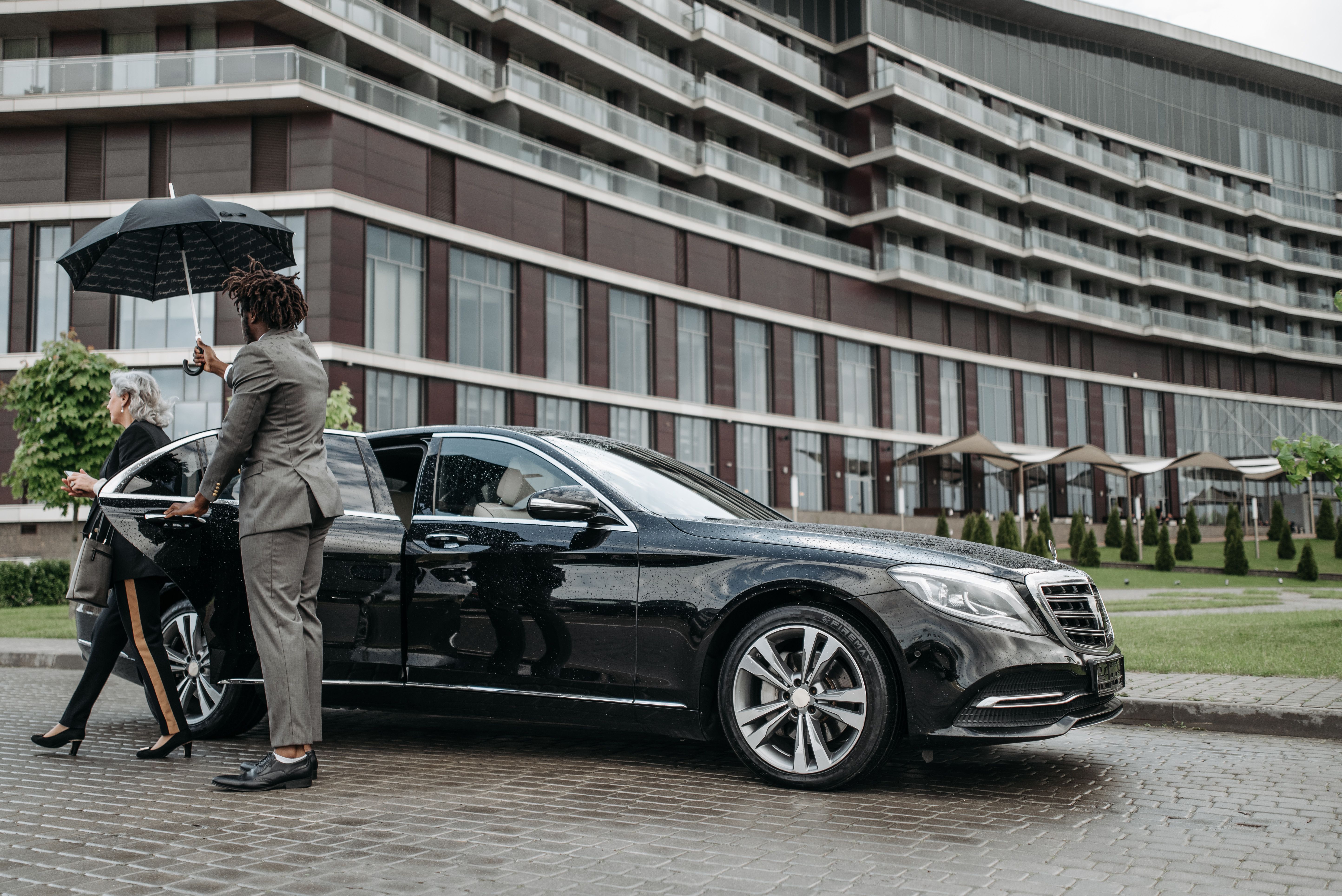 A chauffeur shields a business traveler with an umbrella while she exits a luxury car, illustrating private and attentive service.